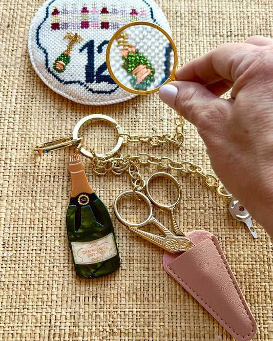 Champagne Tool Chain on grasscloth: gold chain with a champagne bottle charm, embroidery scissors in a blush leather sheath and a magnifier held over a stitched ornament