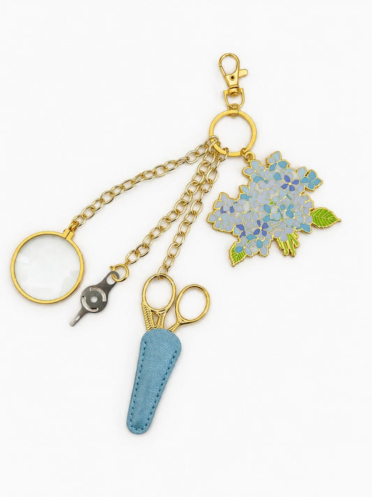 Hydrangea Bloom Tool Chain: Sara Fitz hydrangea enamel charm, gold stork scissors in a powder-blue leather sheath, needle threader, and 5x magnifier on a gold clasp