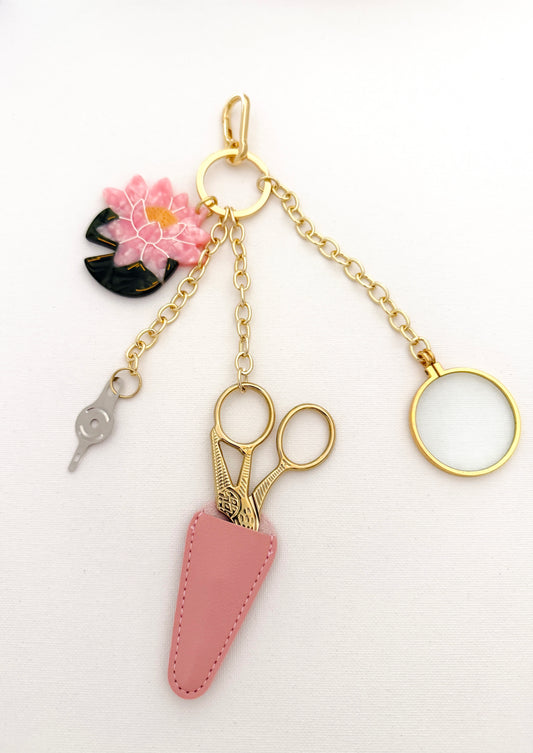 Gold stork scissors in a pink sheath, a folding 5x magnifier and a needle threader on gold chains with a hand-painted pink Japanese water lily charm