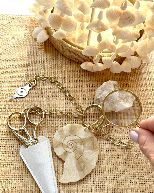 Nautilus Tool Chain on grasscloth under a shell lamp: gold chain, nautilus charm, scissors in a cream leather sheath, needle threader and a magnifier held over a stone