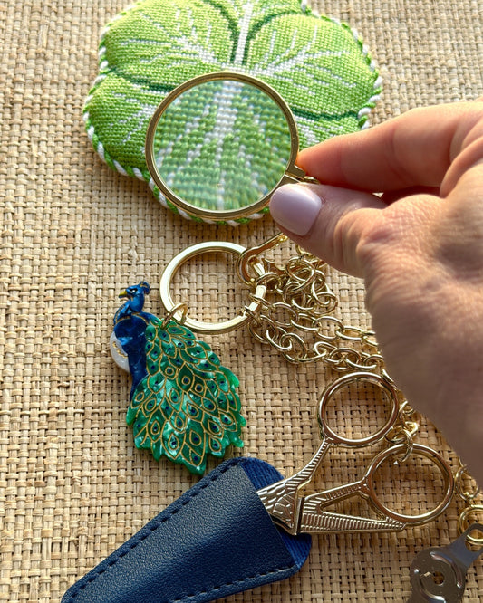 Peacock Tool Chain on grasscloth: peacock charm, gold embroidery scissors in a navy leather sheath, and the magnifier held over a stitched piece