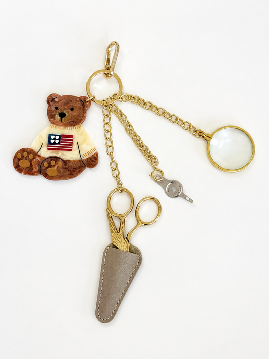 Sweater Bear Tool Chain: hand-painted enamel bear charm in a flag sweater, gold stork scissors in a taupe leather sheath, needle threader, and 5x magnifier on a gold clasp